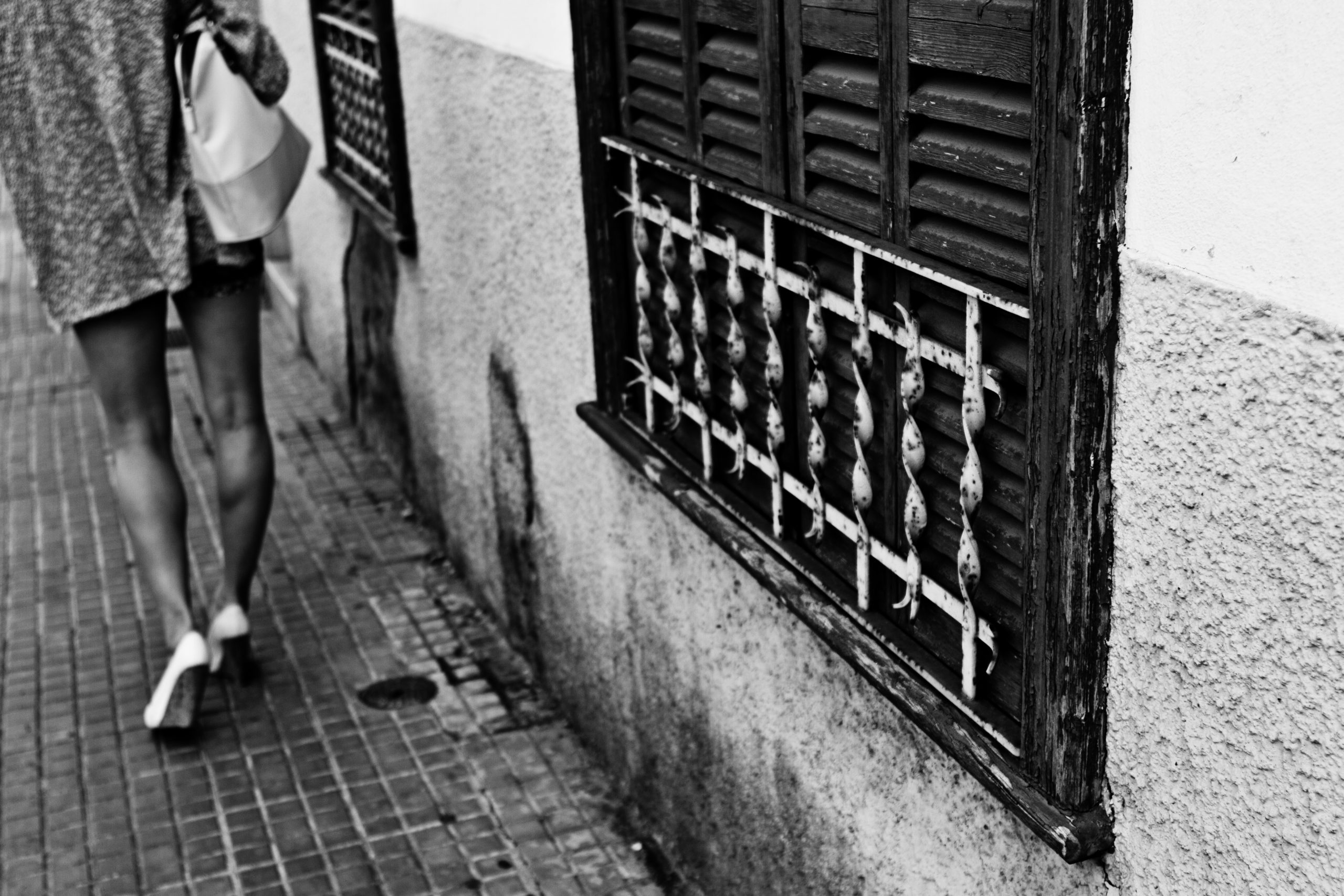 A woman walking past a wooden window with twisted white bars on a dirty street